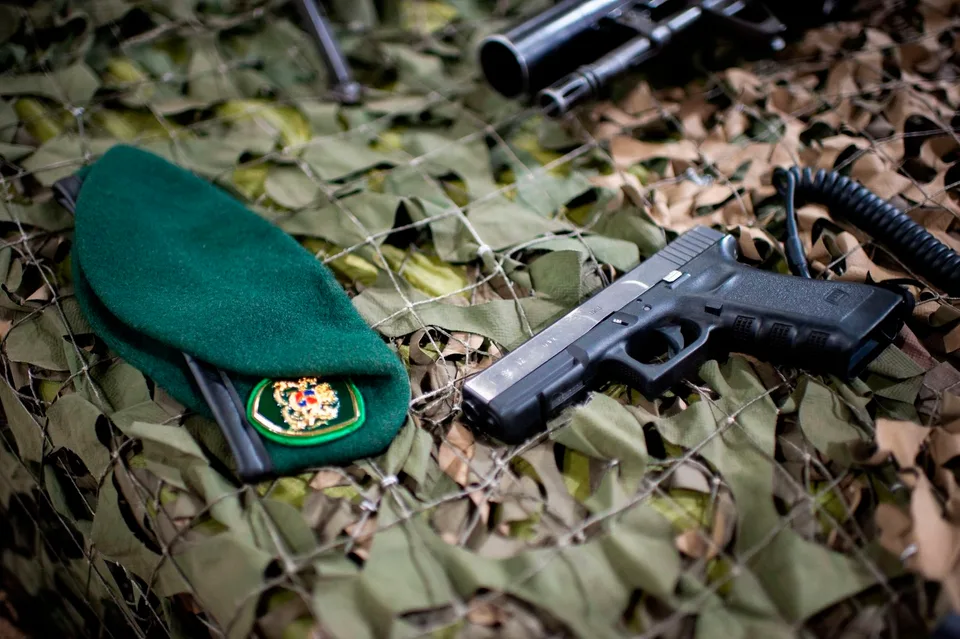 Pistola semiautomática negra sobre un tejido de camuflaje, junto a una boina verde y otras armas parcialmente visibles.
