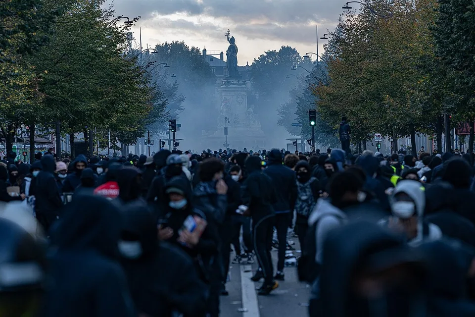 Fotografía documental de los disturbios del 17 de octubre de 2026 en la plaza de la República de París, con manifestantes y humo de gases lacrimógenos.