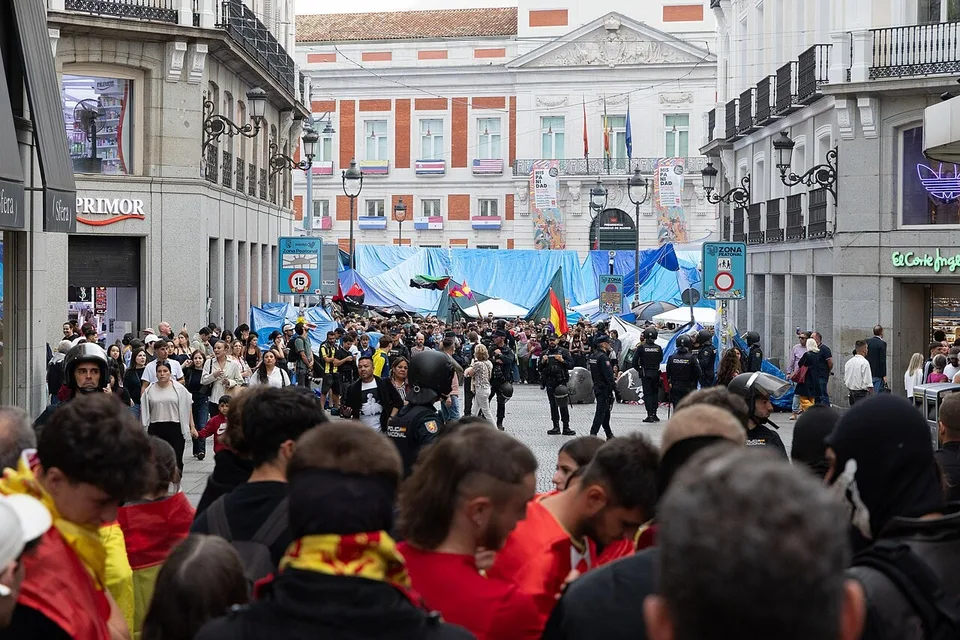 Policías antidisturbios frente a asistentes con banderas españolas en la Puerta del Sol el 12 de octubre de 2026; tiendas de la acampada visibles al fondo.