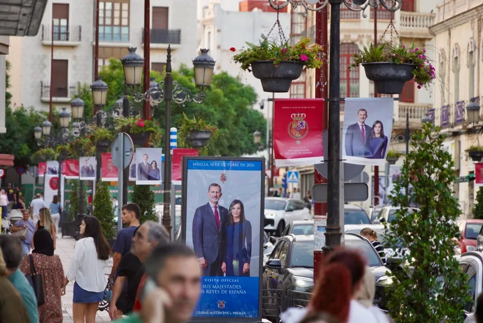 Una calle con farolas y carteles institucionales con retratos de los Reyes, decorada para una visita oficial.