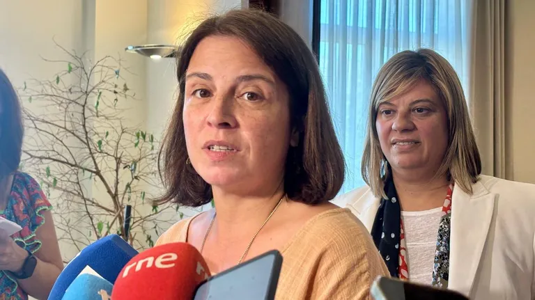 Una mujer habla ante micrófonos de prensa durante una comparecencia, con otra mujer situada detrás.