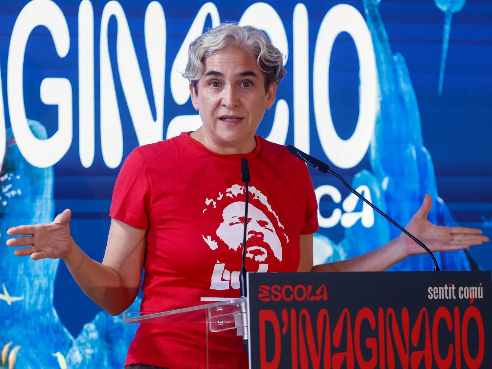 Una mujer de cabello corto y camiseta roja interviene ante un micrófono sobre un escenario político.
